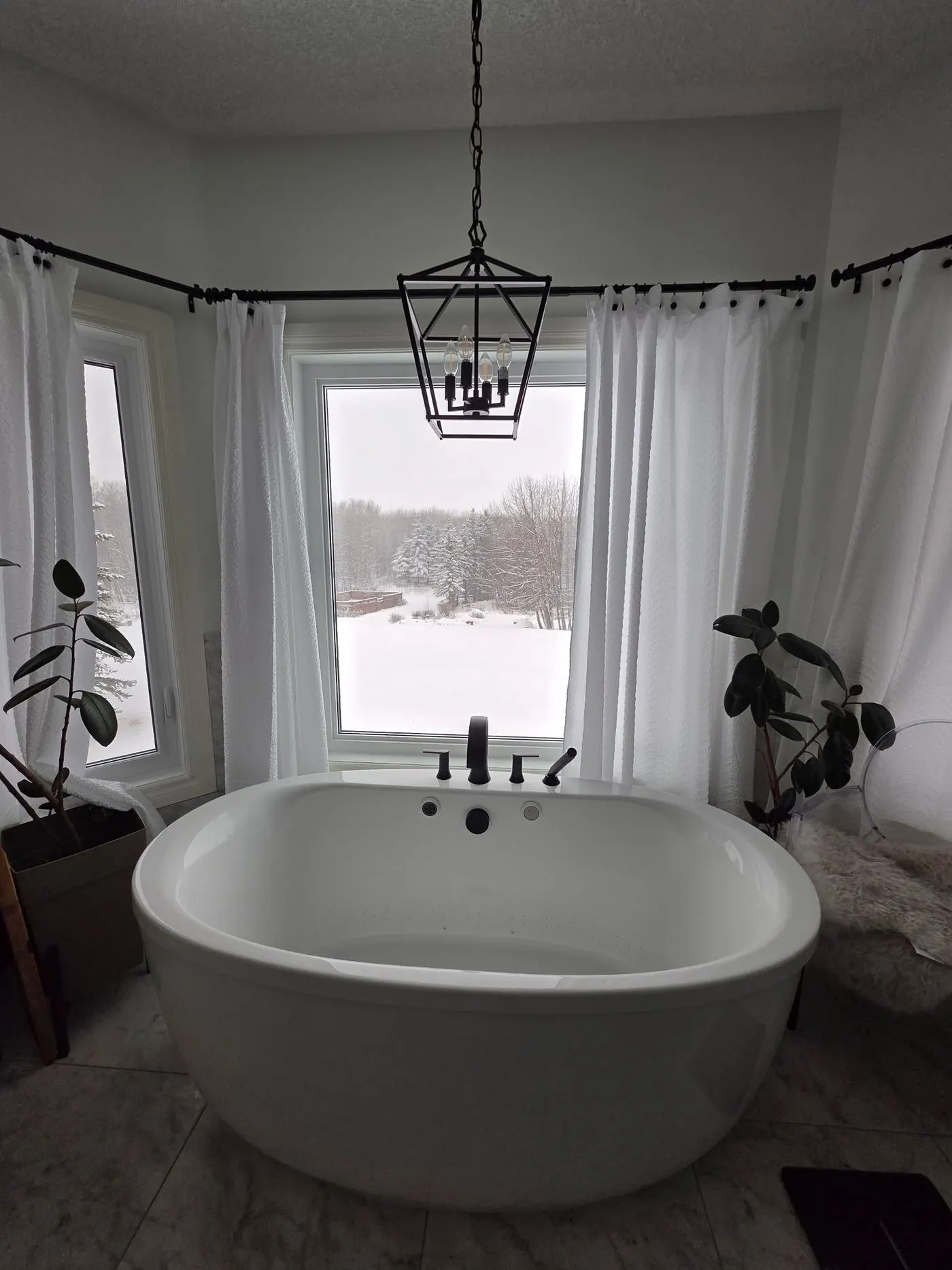 Freestanding soaker tub with a black floor-mounted filler under a lantern pendant, in front of a bright window