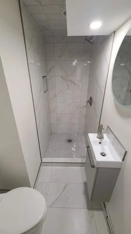 Narrow bathroom with a tiled glass-enclosed shower, wall-hung sink, round mirror and light floor tile
