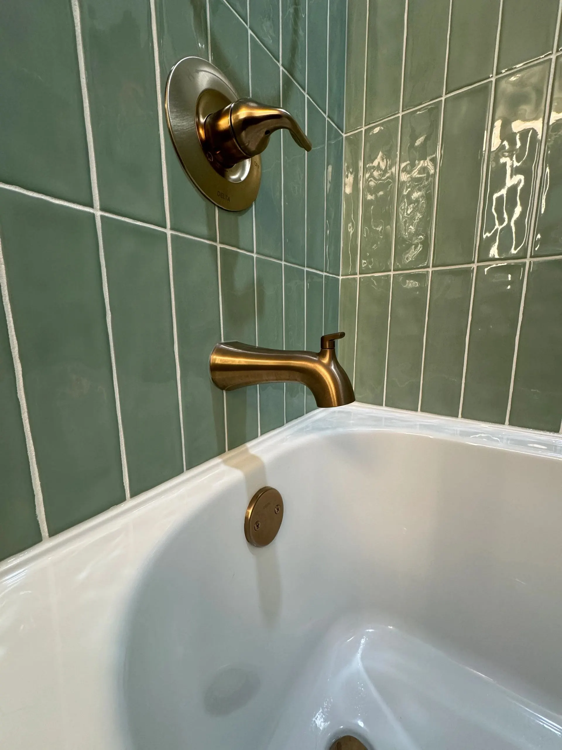 Glossy sage green vertical tile around a white bathtub with a brushed gold spout and drain