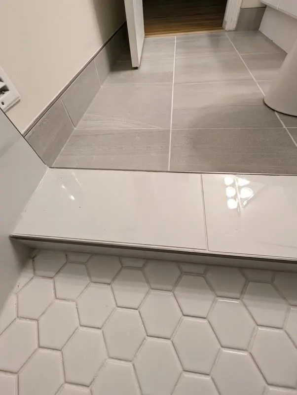 Tiled tub deck with a metal edge trim next to white hexagon floor tile and large grey floor tile