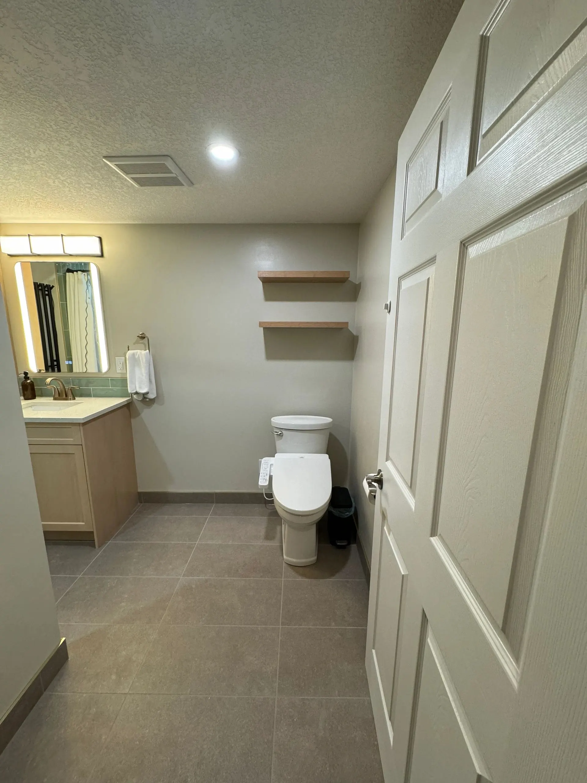 View from the doorway of a bathroom with a light wood vanity, lit mirror, floating wood shelves and grey floor tile