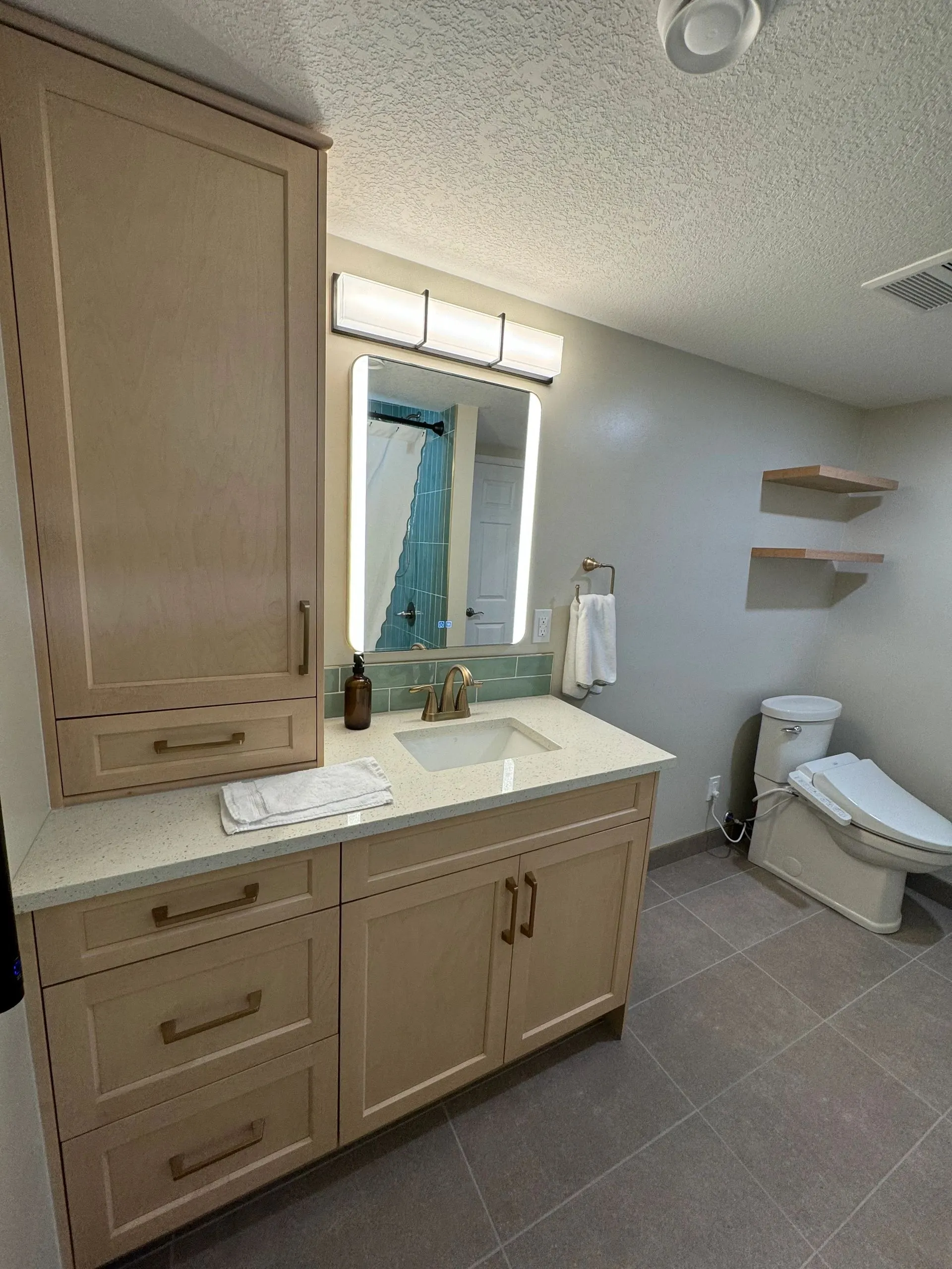 Bathroom with a light wood shaker vanity, brass pulls, quartz top, lit mirror and large grey floor tile